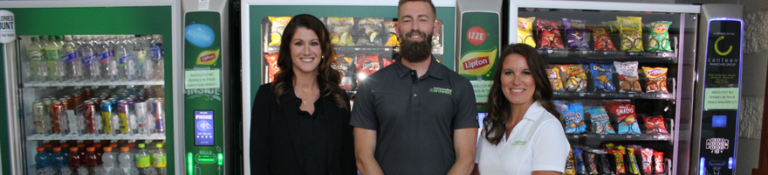 Ashilyn Sunderman (woman) stands on left with two team members (one man in the middle and woman on the right) in front of a line of vending machines and smart coolers.
