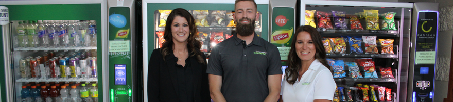 Ashilyn Sunderman (woman) stands on left with two team members (one man in the middle and woman on the right) in front of a line of vending machines and smart coolers.