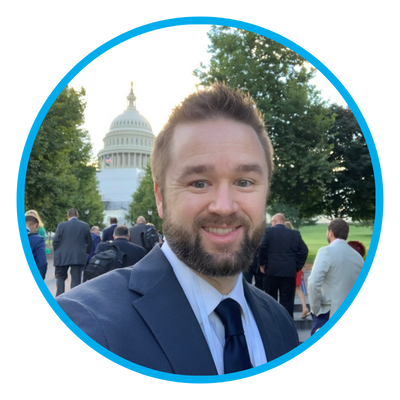 selfie of a man smiling, wearing a suit with the U.S. Capitol behind him.