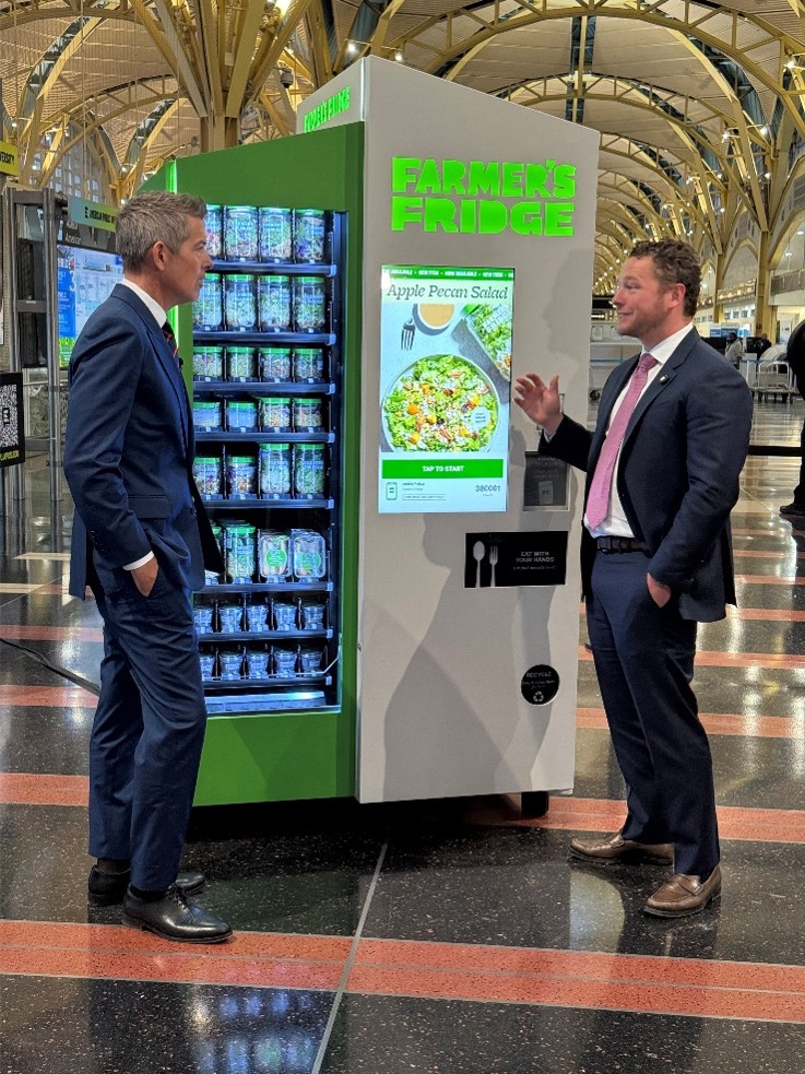 Secretary Duffy and Luke Saunders, Farmer's Fridge Founder and CEO stand talking in front of a Farmer's Fridge vending machine.