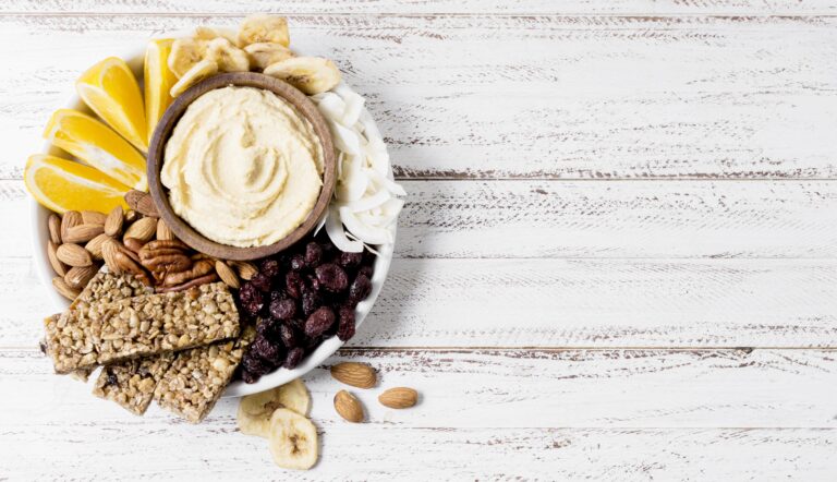 variety of protein snacks all on a plate such as nuts, protein bars, and hummus.